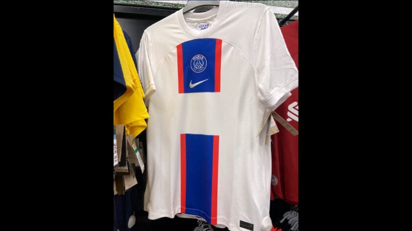 PSG - A nova terceira camisa do PSG para a temporada é predominantemente branca e traz uma faixa azul central com bordas vermelhas, em um padrão “Hechter” invertido, assim como ocorre no manto titular.