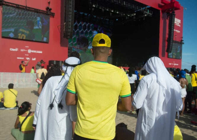 Duas pessoas com trajes típicos da cultura árabe se juntam ao torcedor brasileiro para assistir à partida. 